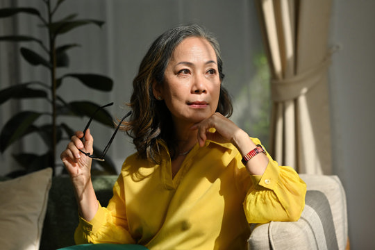 An Asian woman wearing a yellow shirt is seated comfortably on a couch, exuding a relaxed and cheerful demeanor.