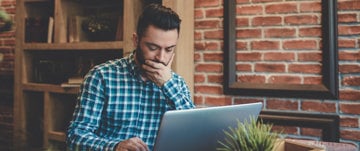 a man in a shirt works at a laptop and holds his hand over his mouth