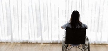 A woman in a wheelchair observes the view outside the window, lost in contemplation.