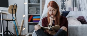 A woman sits on a bed, holding a book, reflecting on her journey with migraines in six impactful words.