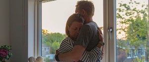 A couple shares a warm hug in front of a window, symbolizing love and intimacy in a serene setting.