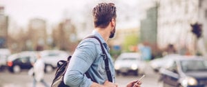 A man carrying a backpack and holding a cellphone walks along a city street, surrounded by urban scenery.