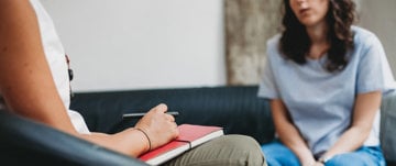 A woman with a notebook sits beside another woman on a couch, sharing thoughts on psychotherapy post-heart attack survival.