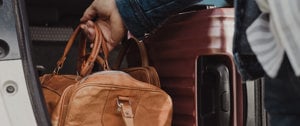 a hand holds a brown leather travel bag in the trunk of a car