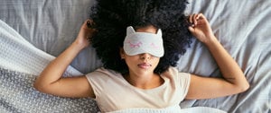 A woman in bed wearing a sleep mask, seeking relief from migraine and sensory overload.