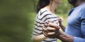 A man and woman hold hands while walking together in a serene park setting, surrounded by greenery.