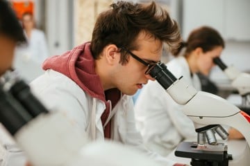 A male student in a lab coat examines a specimen through a microscope in a laboratory setting.