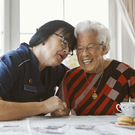 A woman and an older woman share a cup of tea at a table, symbolizing connection and community health support.