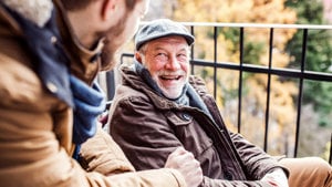 Two men sitting on a balcony, engaged in a lively discussion, surrounded by a serene outdoor atmosphere.